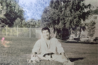 [ai] A man in a karate gi and black belt sitting on the grass, surrounded by trees and a fence in the background. The image is in black and white.
